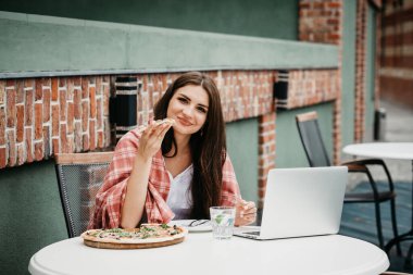 Dizüstü bilgisayar kullanan ve kafe masasında otururken pizza yiyen serbest çalışan genç bir kadın..