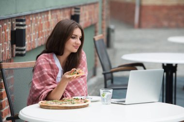 Dizüstü bilgisayar kullanan ve kafe masasında otururken pizza yiyen serbest çalışan genç bir kadın..