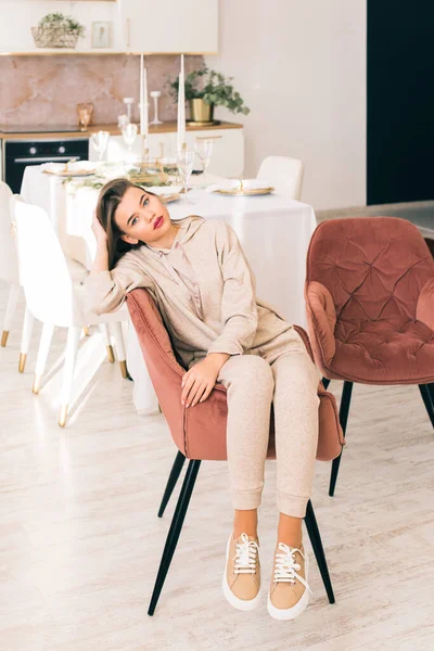 Foto de una mujer en traje deportivo beige y elegantes zapatillas de cuero posando en la silla en una cocina. enfoque selectivo —  Fotos de Stock