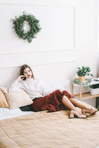 Portrait de femmes à la mode en jupe rouge, chemisier blanc et élégant beige chaussures à talons hauts avec une boucle de chaîne posant dans un lit — Photo