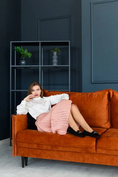 穿着米色短裙、白衬衫和时髦米色黑色休闲鞋、头戴扣子、躺在橙色沙发上的时髦年轻女子的画像. — 图库照片