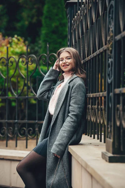 Giovane donna alla moda e bella sorridente 16-20 anni, vestita con un maglione a maglia rosa, occhiali, gonna scura e un classico trench grigio. — Foto Stock