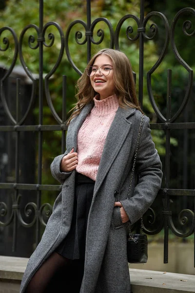 Jeune femme souriante à la mode et belle de 16 à 20 ans, vêtue d'un pull en tricot rose, de lunettes, d'une jupe foncée et d'un trench coat classique gris. — Photo