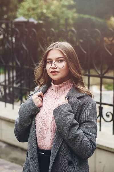 Mujer joven de moda y hermosa 16-20 años, vestida con un suéter de punto rosa, gafas, falda oscura y una gabardina clásica gris. — Foto de Stock