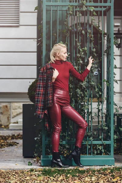 Модная блондинка с красной помадой, позирующая на открытом воздухе. Одетые в красные кожаные леггинсы, водолазку и клетчатую куртку. подходящая фигура — стоковое фото
