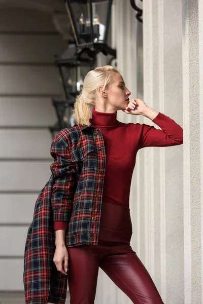 Una hermosa, moderna, chica rubia de moda con un lápiz labial rojo posando al aire libre. Vestido con leggins de cuero rojo, cuello alto y chaqueta a cuadros. figura de ajuste —  Fotos de Stock