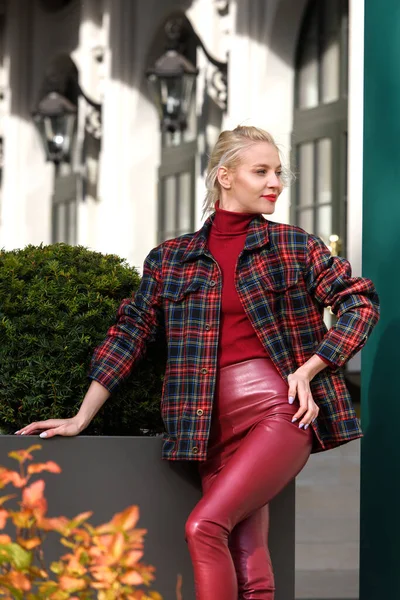 Modisches blondes Mädchen mit rotem Lippenstift, das draußen posiert. Bekleidet mit einer roten Lederleggings, Rollkragen und karierter Jacke. fitte Figur — Stockfoto