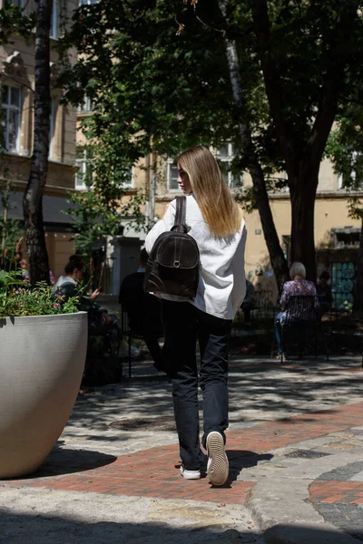 Woman with a brown leather backpack with antique and retro look. Outdoors photo. Woman wearing white jacket and black jeans