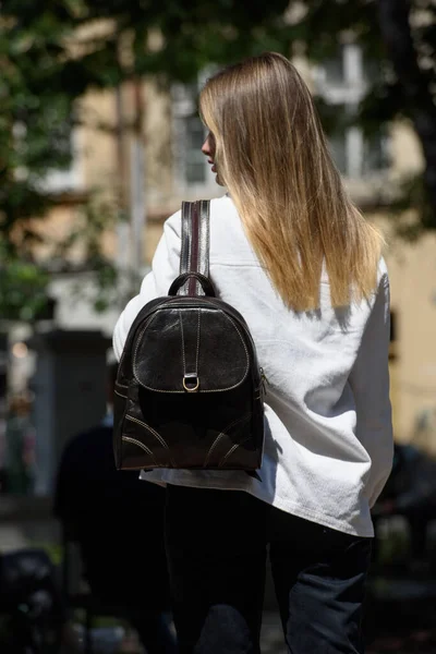 Woman with a brown leather backpack with antique and retro look. Outdoors photo. Woman wearing white jacket and black jeans