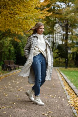 European elegant young woman in a stylish trench coat in a white sweater, blue jeans and white boots walking in a park. warm autumn day.