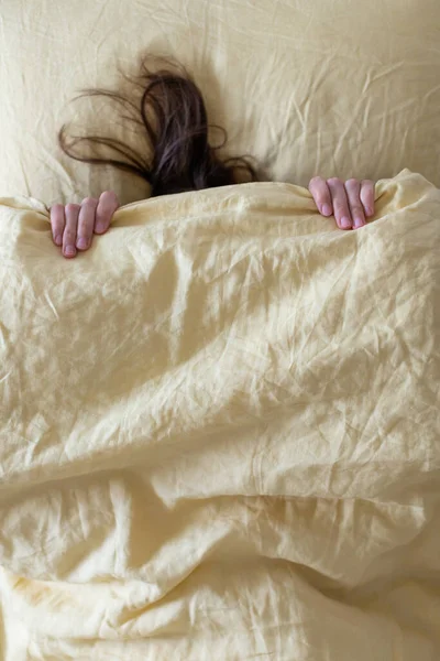 침대에 있는 여자는 머리에 담요를 덮고 있고, 그녀의 손 과 머리카락을 볼 수있습니다. 겨울을 피해 숨는다는 개념 은. — 스톡 사진