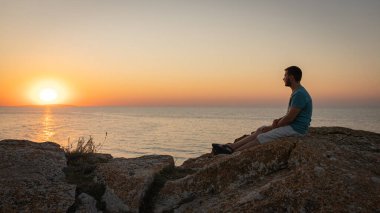 Bir adam gün batımını seyretmekten hoşlanır, bir kayanın üzerinde oturarak..