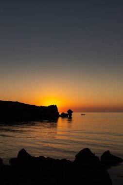 Denizde gün batımı, kayalar ve güneş, siluet ve balıkçılarla dolu tekne.