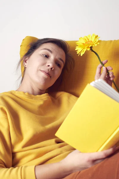 Güzel bir kız, elinde bir kitap ve çiçekle sarı bir sandalyede oturmayı hayal eder.. — Stok fotoğraf