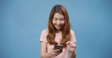 Genç Asyalı bayan telefonu olumlu bir ifadeyle kullanıyor, geniş bir şekilde gülümsüyor, gündelik giysiler içinde mutluluğu hissediyor ve mavi arka planda tek başına duruyor. Mutlu, mutlu, mutlu kadın başarıya sevinir..