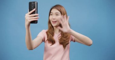 Gülümseyen, sevimli Asyalı bir kadın akıllı telefonuyla fotoğraf çekiyor. Günlük kıyafetlerle pozitif bir ifade takınıyor ve mavi arka planda tek başına duruyor. Mutlu, mutlu, mutlu kadın başarıya sevinir..