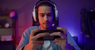 Mutlu Asyalı oyuncu kulaklık yarışması takıyor ve modern evdeki oturma odasında akıllı telefonlu neon ışıklı video oyunu oynuyor. Esport oyun çevrimiçi, ev karantina etkinliği.