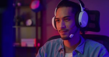 Mutlu Asyalı oyuncu kulaklık takıyor ve gece modern evdeki oturma odasında renkli ışıklı bilgisayar oyunu oynuyor. Esport akış oyunu çevrimiçi, ev karantina etkinliği kavramı.