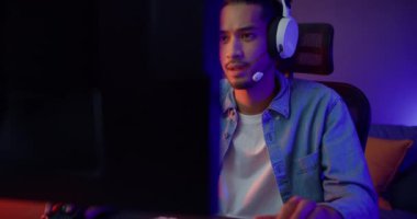 Mutlu Asyalı oyuncu kulaklık takıyor ve gece modern evdeki oturma odasında renkli ışıklı bilgisayar oyunu oynuyor. Esport akış oyunu çevrimiçi, ev karantina etkinliği kavramı.