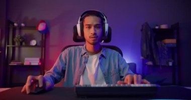 Mutlu Asyalı oyuncu kulaklık takıyor ve gece modern evdeki oturma odasında renkli ışıklı bilgisayar oyunu oynuyor. Esport akış oyunu çevrimiçi, ev karantina etkinliği kavramı.