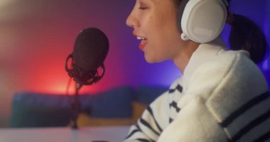 Mutlu Asyalı bayan blogger müzik etkeni bilgisayarda kulaklıklı bir podcast ve oturma odasındaki ev stüdyosunda izleyicilerle mikrofon konuşması kaydediyor. Evde kal, içerik yaratıcısı konsepti.