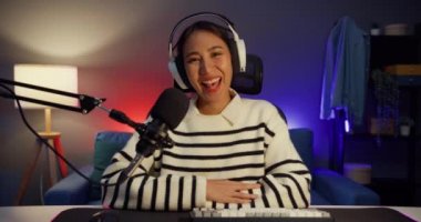Mutlu Asyalı bayan blogger müzik etkilici kamera yayınına bakıyor kulaklık takıyor mikrofondan canlı yayında dinleyicilerle oturma odasındaki stüdyoda konuşuyorlar. İçerik oluşturucu kavramı.