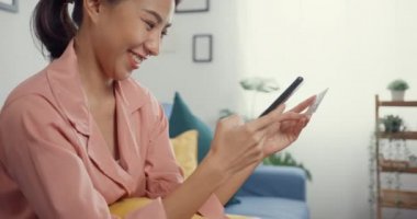 Genç Asyalı mutlu bayan cep telefonu siparişini online alışveriş ürünü olarak kullanıyor ve kredi kartıyla ödeme yapıyor. Oturma odasındaki kanepede oturuyor. Karantina aktivitesi, koronavirüsü önlemek için eğlenceli aktivite.