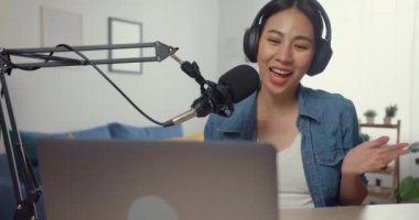 Mutlu Asyalı kız dizüstü bilgisayarda kulaklıklı bir podcast ve oturma odasında izleyicilerle mikrofon konuşması kaydediyor. Kadın podcaster kendi stüdyosundan ses yayını yapıyor. Ev konseptinde kal..