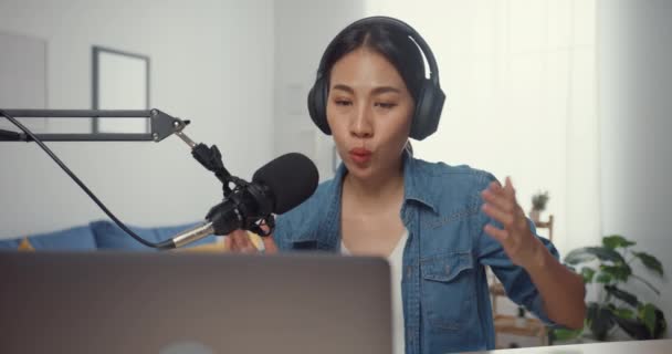 幸せなアジアの女の子は リビングルームで聴衆とヘッドフォンやマイクトークとラップトップコンピュータ上のポッドキャストを記録します 女性のポッドキャストは 彼女のホームスタジオからオーディオポッドキャストを作ります 家のコンセプトにとどまる — ストック動画