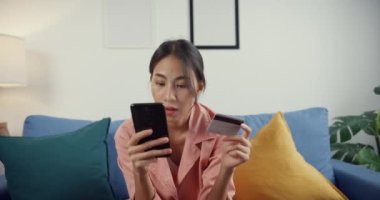 Kapalı alan Asyalı genç bayan cep telefonu siparişini online alışveriş ürünü olarak kullanıyor ve kredi kartıyla fatura ödüyor evdeki oturma odasında koltukta oturuyor, karantina aktivitesi, koronavirüs önleme için eğlenceli etkinlik.