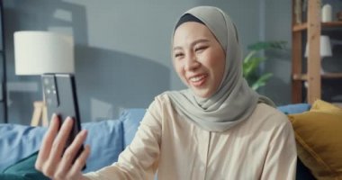 Çekici, neşeli, genç, Asya 'lı, tesettürlü, Müslüman, gündelik bir şekilde kanepede oturan güzel bir kadın. Oturma odasındaki arkadaşınla akıllı telefon görüşmeleri yap. Kız İslami yaşam tarzı kavramı.