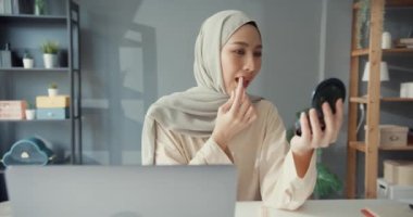 Çekici, neşeli, Asyalı, genç, Müslüman iş kadını. Laptopuyla tesettüre oturmuş evinde dudaklarına ruj sürüyor. Kız İslami yaşam tarzı kavramı.