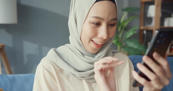 ソファの上にカジュアルな座ってヒジャーブの魅力的な陽気な若いアジアのイスラム教徒の美しさの女性は自宅で居心地の良いリビングルームでオンラインショッピングをお楽しみください 女の子イスラム隔離ライフスタイルコンセプト — ストック動画