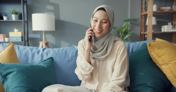 自宅の居心地の良いリビングルームで友人や家族とのカジュアルな使用電話でヒジャーブ州の魅力的な陽気な若いアジアのイスラム教徒の美しさの女性 女の子イスラム社会的距離隔離ライフスタイルコンセプト — ストック動画