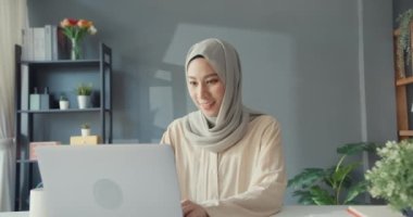 Çekici, neşeli, Asyalı, genç, Müslüman iş kadını, gündelik olarak masada oturuyor. Evdeki oturma odasında çalışan dizüstü bilgisayarı kullanıyor. İnsanlar dini İslam yaşam tarzı konsepti.