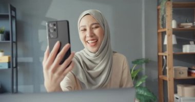 Çekici, neşeli, Asyalı, genç, Müslüman iş kadını, gündelik olarak masa başında oturan, evinde oturan bir arkadaşınla akıllı telefon konferansını kullan. Kız dini İslam yaşam tarzı konsepti.
