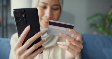 Yakın plan Çekici, genç, Asyalı, İslamcı, tesettürlü güzel kız, gündelik bir şekilde kanepede oturuyor. Telefonu kullan. Başarılı online alışverişin tadını çıkar..