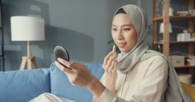 Çekici, neşeli, Asyalı, genç, Müslüman, tesettürlü güzel bir kadın, rahat bir oturma odasında dudaklarına ruj sürmekten zevk alıyor. Kız İslami güzellik yaşam tarzı konsepti.