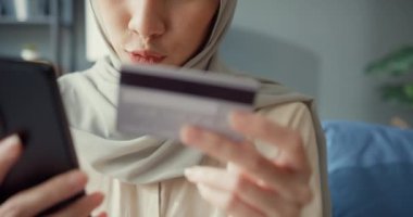 Yakın plan Çekici, genç, Asyalı, İslamcı, tesettürlü güzel kız, gündelik bir şekilde kanepede oturuyor. Telefonu kullan. Başarılı online alışverişin tadını çıkar..
