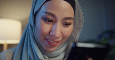 Yakın plan, çekici, Asyalı, genç, neşeli, Müslüman bir kadın, gündelik bir şekilde kanepede oturuyor ve telefon kullanıyor. Geceleri evdeki oturma odasında online alışverişin tadını çıkarıyor. Kız İslami yaşam tarzı kavramı.