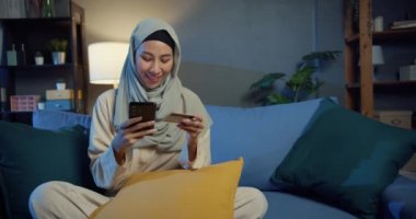 Çekici, neşeli, genç, Asya 'lı Müslüman, tesettürlü, sıradan gülüşlü, telefon kullanan, rahat bir oturma odasında kredi kartıyla elektronik alışveriş yapan güzel bir kız..