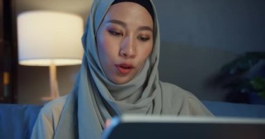 Yakın plan, çekici, Asyalı, genç, neşeli, Müslüman kadın, gündelik tesettürde oturuyor, geceleri evdeki rahat oturma odasında dijital tablet kullanıyor. Kız milleti İslam yaşam tarzı kavramı.