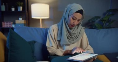 Çekici, neşeli, Asyalı, genç, Müslüman, tesettürlü güzel kadın dizüstü bilgisayarlı koltukta oturuyor ve geceleri evdeki rahat oturma odasında dijital tablet kullanıyor. Kız milleti İslam yaşam tarzı kavramı.