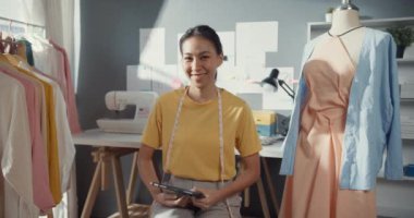 Neşeli profesyonel Asyalı moda tasarımcısı sıradan dijital tabletli gülümseme ile kameraya kendine güvenerek bak. Bayan terzi öğrenci atölyedeki yeni koleksiyonuyla gurur duyuyor..