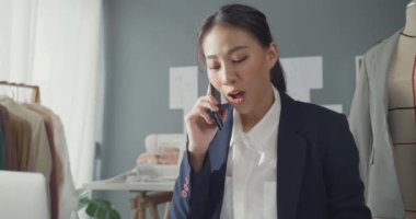 Masada oturan profesyonel neşeli Asyalı iş kadını tasarımcı dizüstü bilgisayarı kullanıyor. Akıllı telefonu kullanarak müşterinin satın alma siparişini stüdyo işyerindeki çevrimiçi pazardan onaylıyor. Başarılı bir iş..