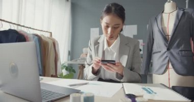 Masada oturan profesyonel neşeli Asyalı iş kadını tasarımcı akıllı telefon satın alma siparişine, stüdyo işyerindeki dizüstü bilgisayarında stok envanteri bulunan online pazar yeri satın alma talimatlarına bakın..