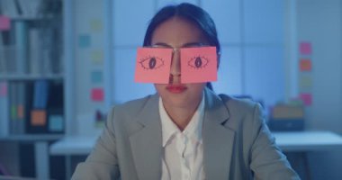 Masada oturan Asyalı genç iş kadını geceleri ofiste dizüstü bilgisayar başında uyuyakalıyor. Yorgun bir kadın. Gözlerinin üzerinde iki post-it var. İş yerinde uyuyan yüz üzerine yapışkan notlar..