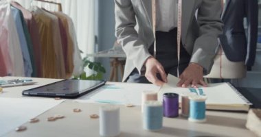 Yakın plan Asyalı iş kadını terzisi taslak defteriyle meşgul. Stüdyoda çalışma masasında dijital tablet, elbise deseni ve dağınık masa tasarımcısı ile karşılaştır..