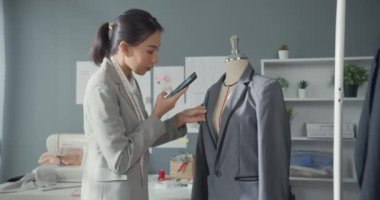 Masada oturan neşeli Asyalı iş kadını moda tasarımcısı akıllı telefon kullan. Stüdyo işyerindeki siparişi doğrulamak için internetten fotoğraf çek. İş kurma.