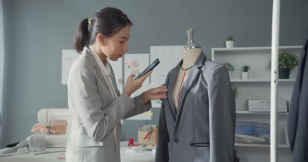 テーブルに座ってスーツコレクションを持つ陽気なアジアのビジネス女性ファッションデザイナーは スタジオの職場での購入注文を確認するためにスマートフォンを使用して写真をオンライン市場を取ります ビジネスを始める — ストック動画